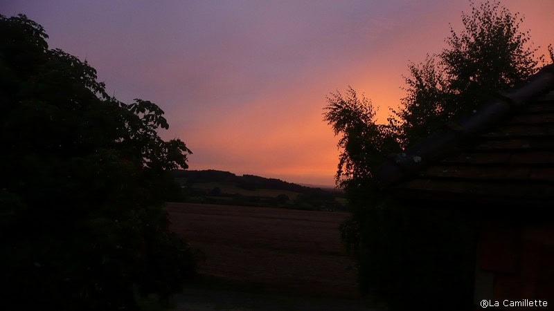 Een rustige zonsondergang boven een landelijk landschap in Frankrijk, met tinten roze en oranje aan de hemel. Silhouetten van bomen omlijsten het serene tafereel aan beide kanten, met een heuvel op de achtergrond en een vredig veld op de voorgrond. Een deel van een dak is zichtbaar aan de rechterkant.