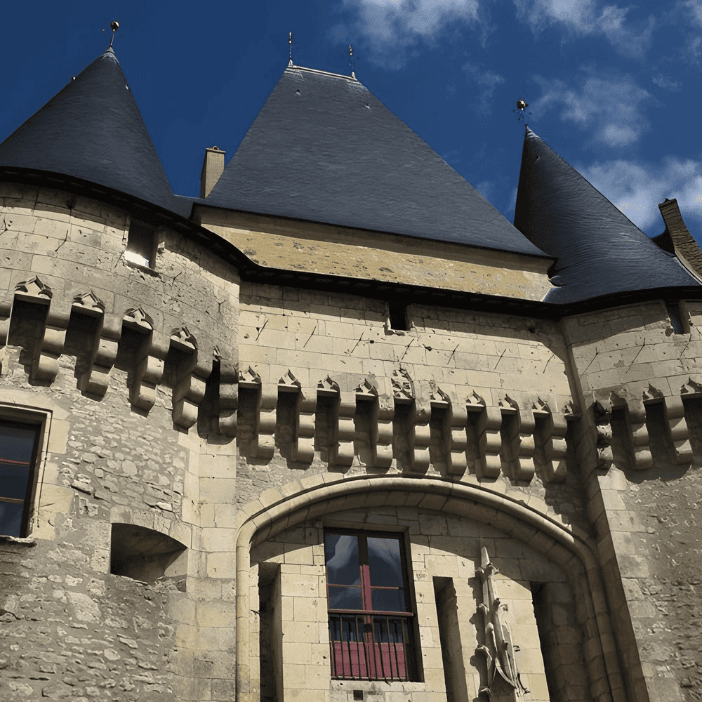 Een historisch stenen gebouw op het Franse platteland heeft puntige, donkere leien daken en decoratieve kantelen onder een blauwe lucht. Een groot boograam met rode lijsten is zichtbaar en kleine houtsnijwerken sieren de stenen gevel, wat de architectonische details benadrukt die kenmerkend zijn voor een gîte.