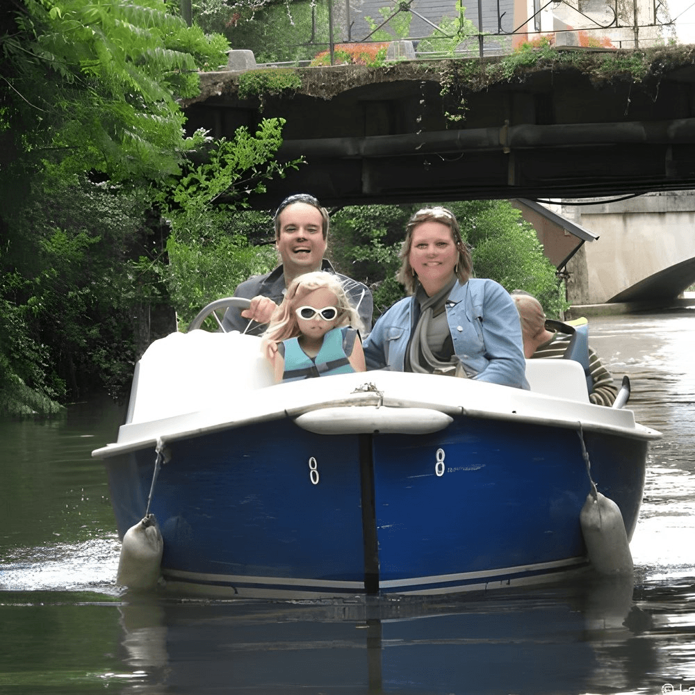 Een gezin geniet van een boottocht door een rustig kanaal. Een jong kind met een zonnebril zit vooraan en lacht, terwijl twee volwassenen achter haar zitten, waarvan er één aan het roer staat. Boven haar hoofd overspant een stenen brug het kanaal. Weelderig groen omringt hen en op de achtergrond is een huis met houten balken te zien, mogelijk een schilderachtig huisje.