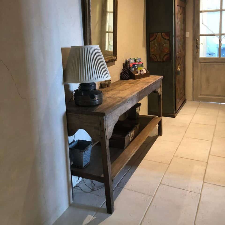 Een hal met een houten consoletafel tegen de muur, met daarboven een geplooide lampenkap, straalt rustieke charme uit die doet denken aan een schilderachtig huisje in Frankrijk. Beneden staat een rieten mand op de onderste plank. Er hangt een spiegel in vintagestijl boven en een decoratieve kast met kleurrijke patronen staat bij de deur met glazen panelen.