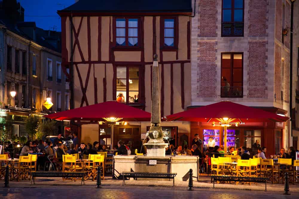 Een levendige avondscène toont een druk openluchtcafé met felrode parasols en felgele stoelen op een geplaveide straat. Diners verzamelen zich rond tafels bij een stenen fontein met ingewikkelde details, terwijl de achtergrond charmante historische architectuur toont, die doet denken aan een schilderachtige gîte-setting.