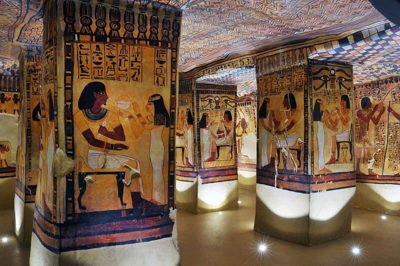 Het interieur van een oud Egyptisch graf toont levendige hiërogliefen en kleurrijke schilderingen op muren en zuilen. Kunstwerken tonen figuren in traditionele kledij, bezig met verschillende activiteiten, met ingewikkelde geometrische patronen op het plafond. Zachte verlichting benadrukt deze historische scène op zachte wijze.
