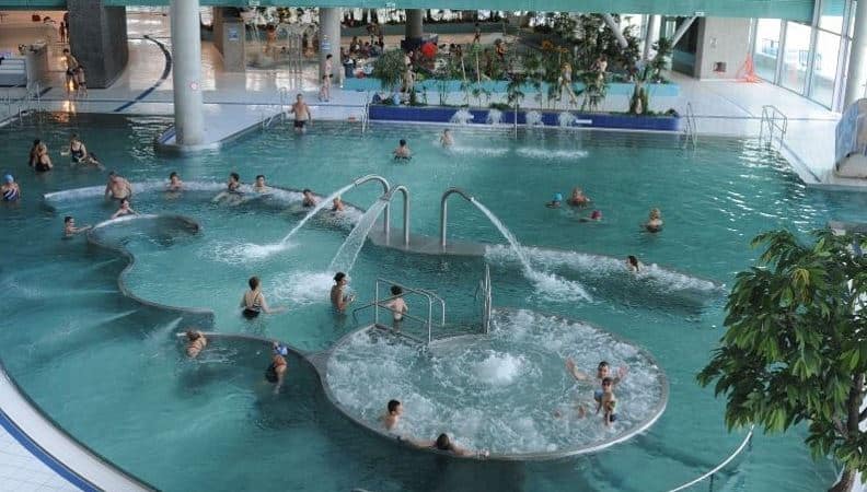 Een overdekt zwembadcomplex met mensen die genieten van verschillende wateractiviteiten, waaronder een cirkelvormig bubbelbad. Rondom het zwembad staan potplanten en een loopbrug die de ontspannen sfeer versterkt. De faciliteiten hebben helder water en uitstekende verlichting, met een vleugje rustieke charme.