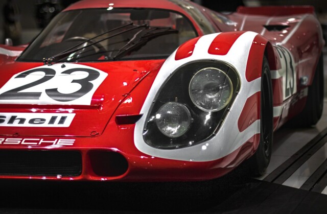 Een rode klassieke raceauto met witte strepen op de motorkap en het nummer 23. Hij heeft ronde koplampen en een strak aerodynamisch design. De voorkant van het voertuig toont prominent het Porsche-logo, wat de essentie van snelheid vastlegt die vaak voorkomt in races zoals die in Frankrijk.