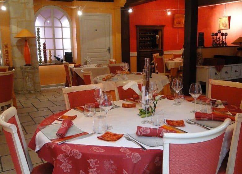 Een warm restaurantinterieur met ronde tafels bedekt met witte en rode tafellakens met bloemenmotief. Elke tafel is gedekt met glazen, servetten en kleine bordjes. Rode en witte stoelen omringen de tafels. De gele en rode muren roepen een charmante Franse plattelandssfeer op met decoratieve kunst en zachte verlichting.