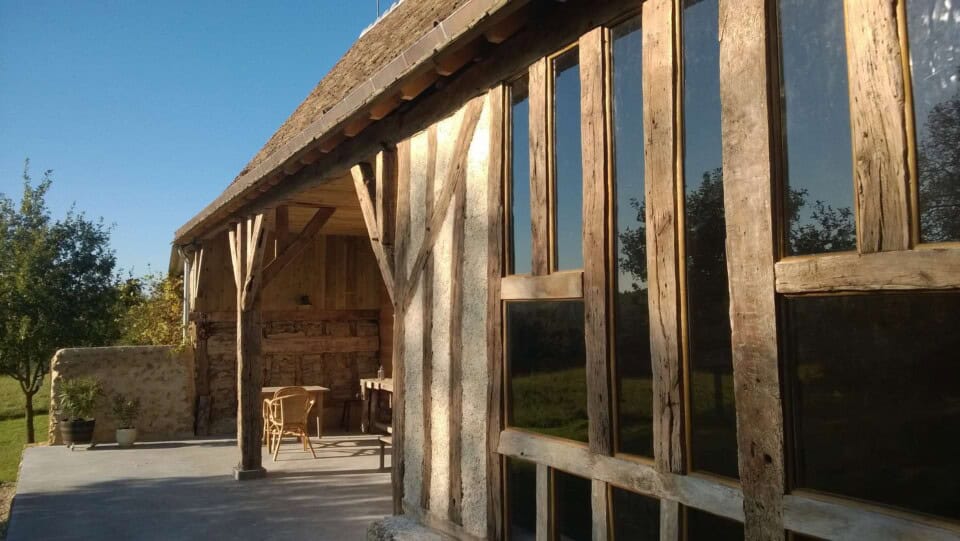 Een rustieke houten hut, die doet denken aan een gîte op het platteland, heeft grote glazen ramen die baden in zonlicht. Houten balken en een schuin dak dragen bij aan de charme. Een patio met rieten stoelen kijkt uit over de weelderige groene natuur, wat de serene, pastorale omgeving onder de helderblauwe lucht versterkt.