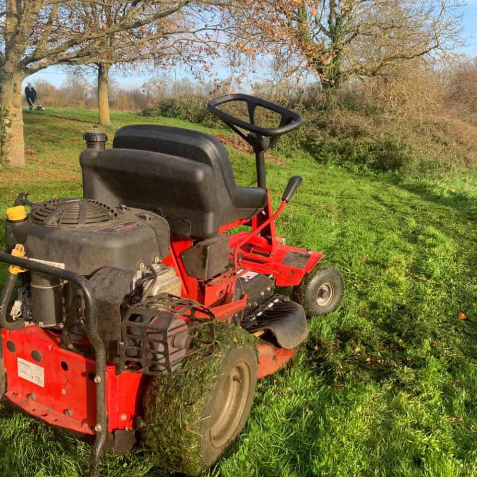 Een rode zitmaaier met een zwarte zitting rust op een stukje weelderig, groen gras. Het zichtbaar gebruikte cirkelvormige maaidek is bestrooid met grasresten. Bladloze bomen steken af tegen de blauwe lucht en bieden een gevoel van roest en sereniteit dat doet denken aan een verblijf in een vredige gîte in de late herfst.